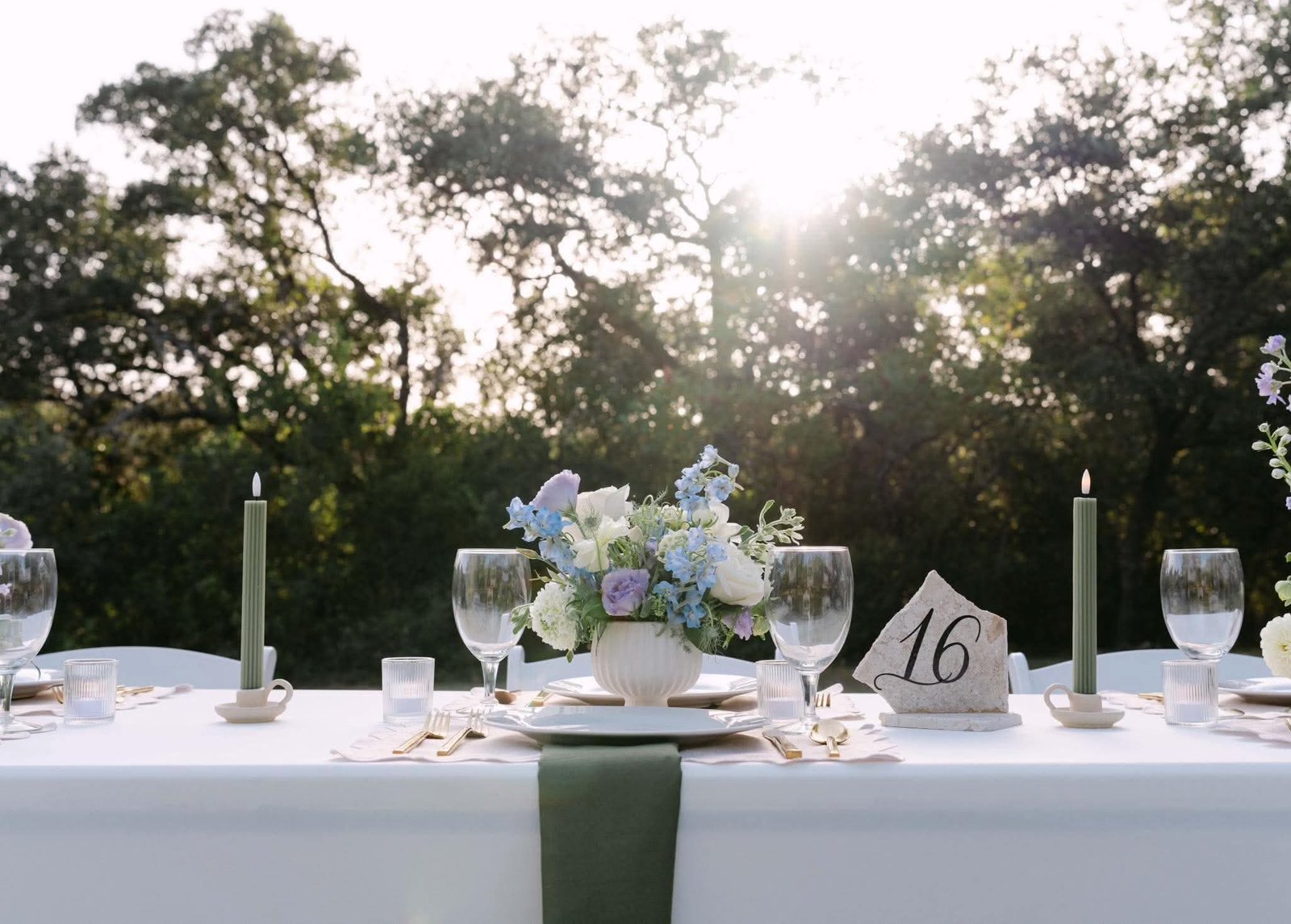 Low white footed vase of blue and lavender blooms between fluted green tapers and a stone table number, backlit at sunset