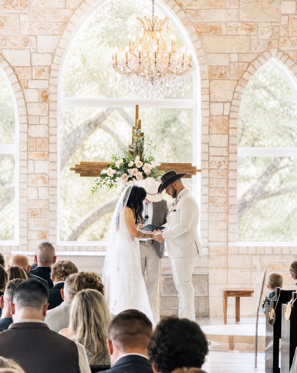 Bride in lace and groom in a cowboy hat exchanging vows in a limestone chapel, a floral wooden cross and crystal chandelier behind them