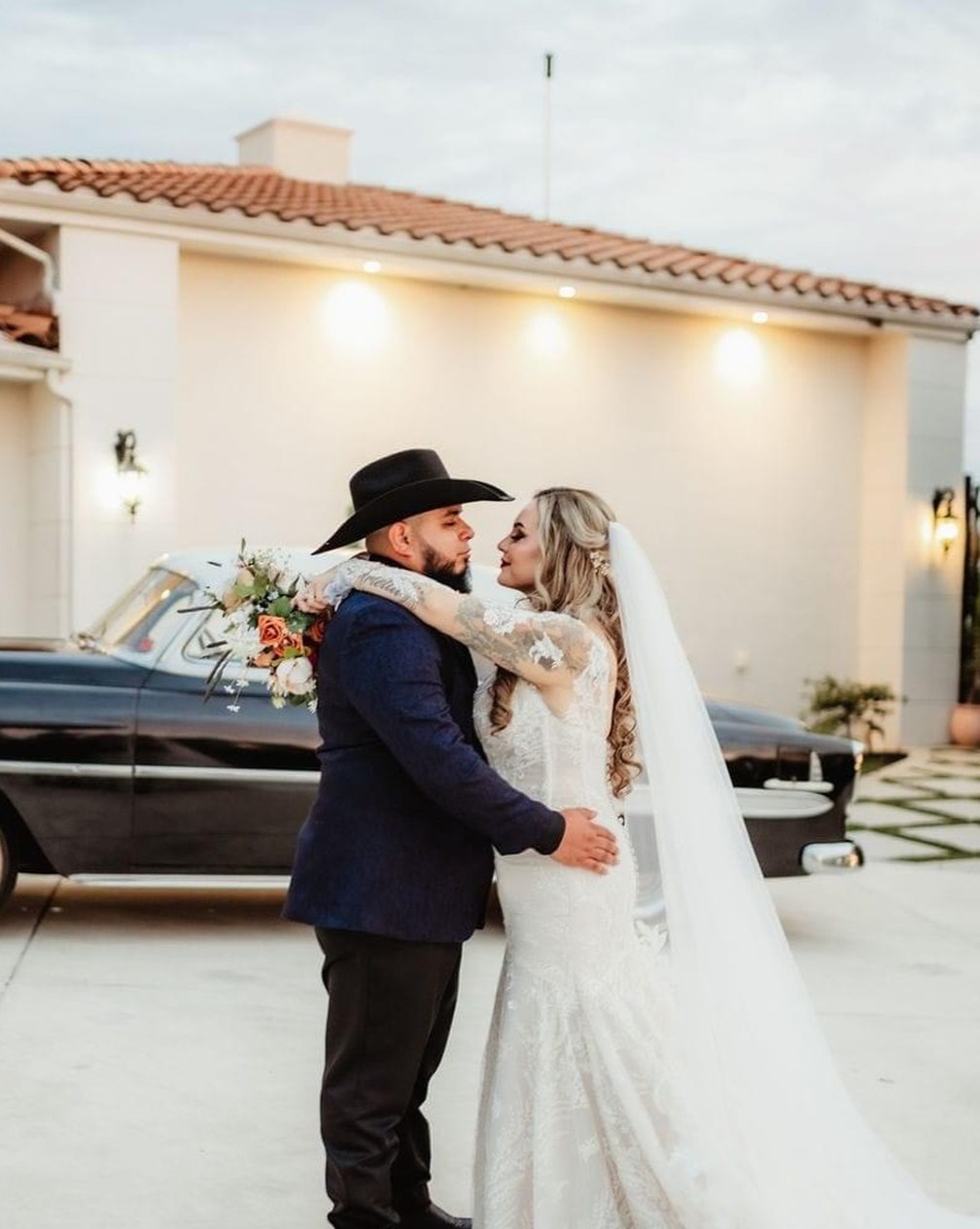 Bride and groom embracing at dusk beside a vintage car, the groom in a cowboy hat