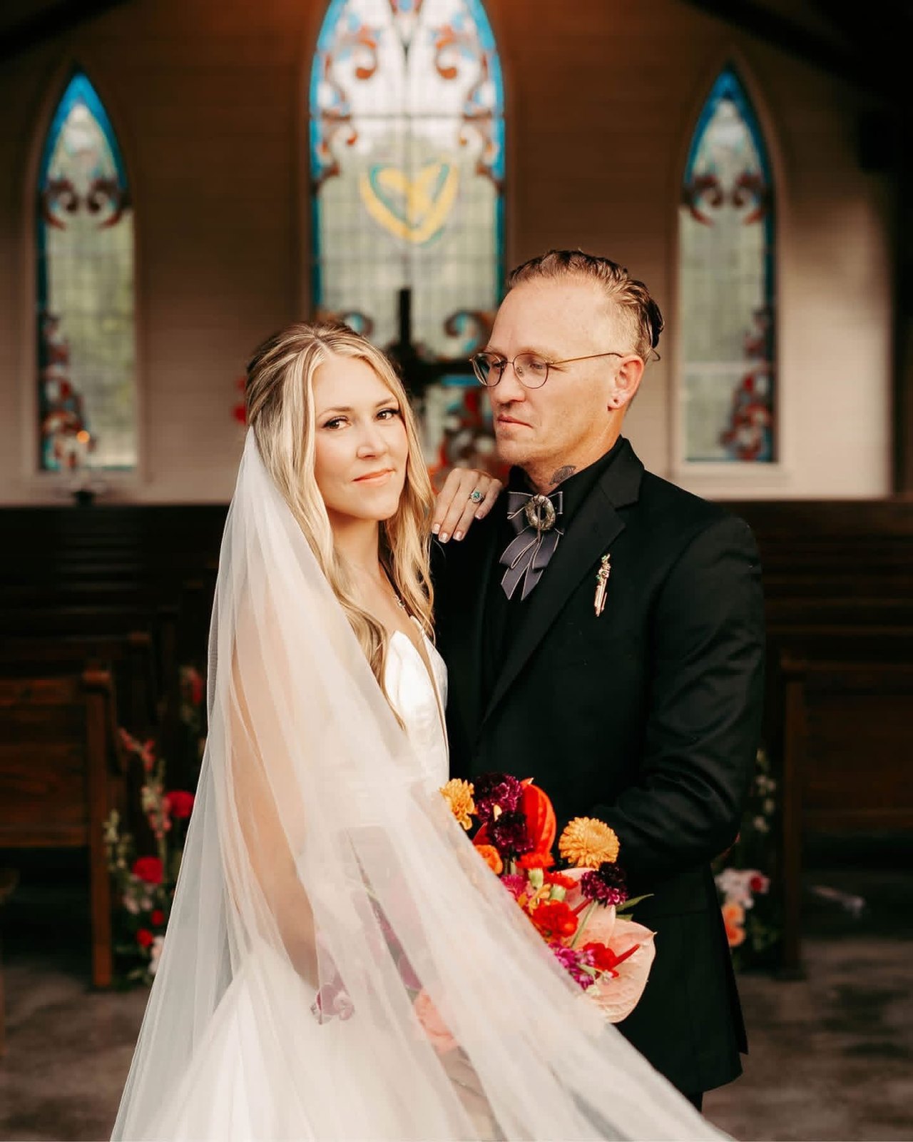 Bride in a cathedral veil and groom in a black suit in a chapel, stained-glass windows behind them and a bouquet of red and orange blooms