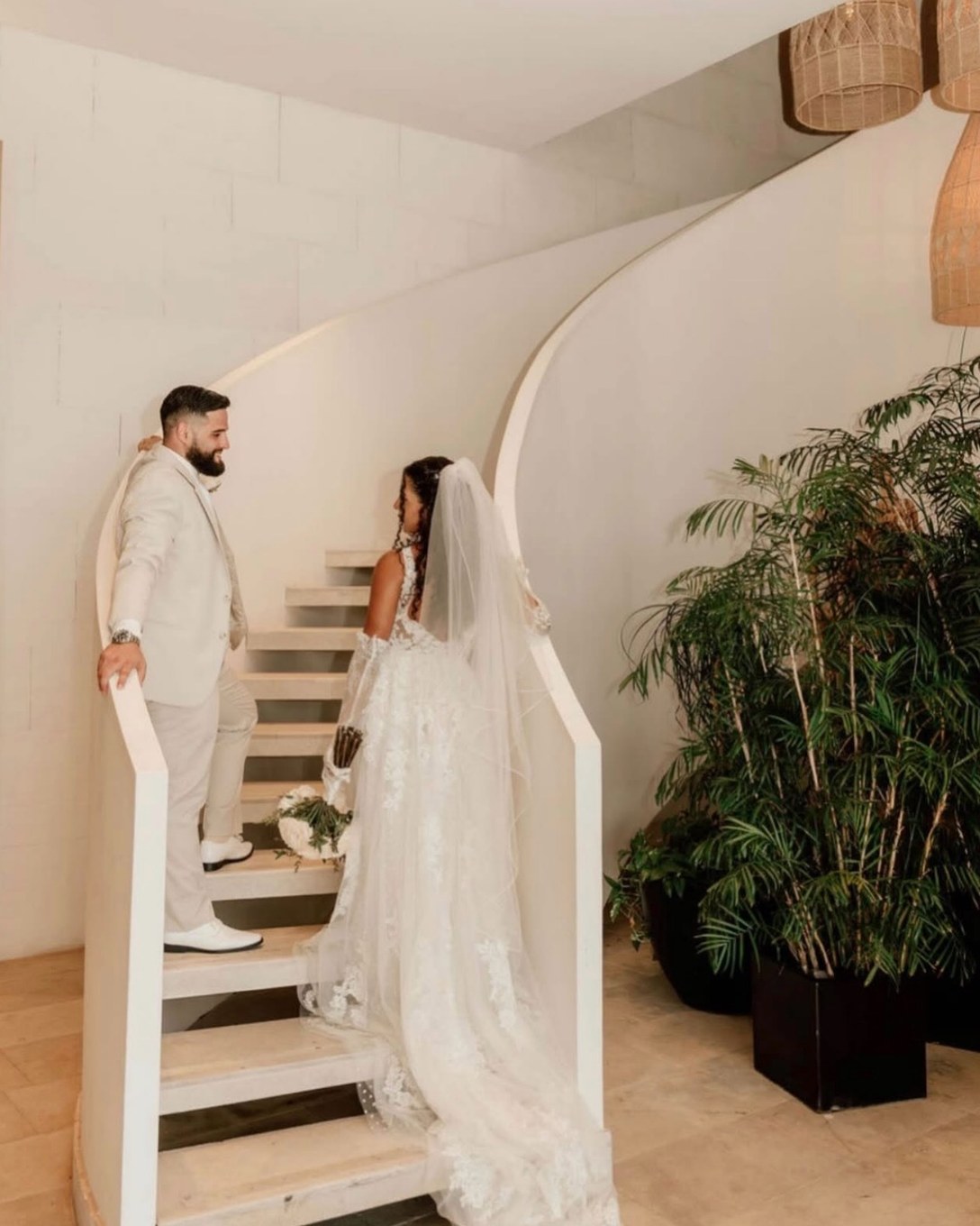 Bride looking up a curved white staircase toward the groom, her lace train on the steps