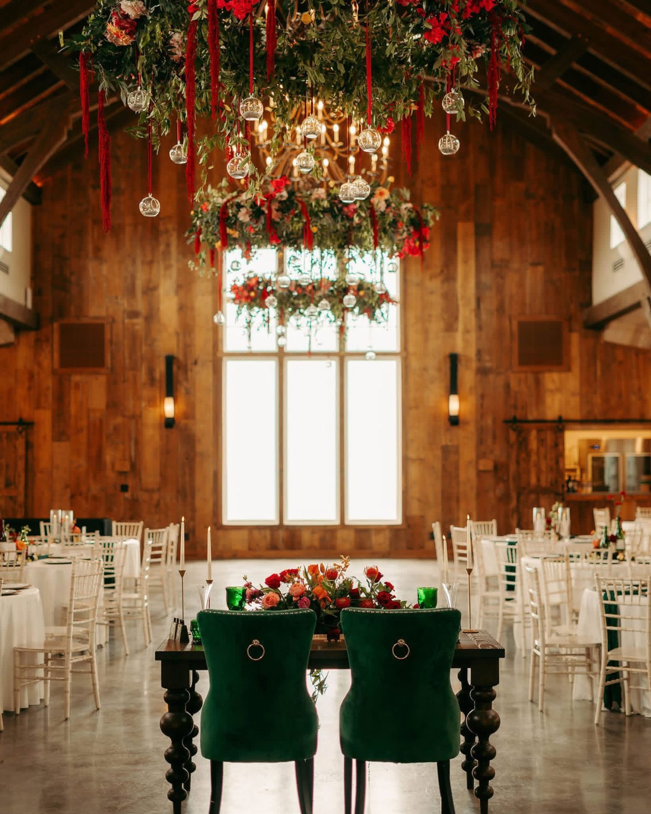 Barn reception with red and green florals hung above the sweetheart table, emerald velvet chairs and round tables set in white linen