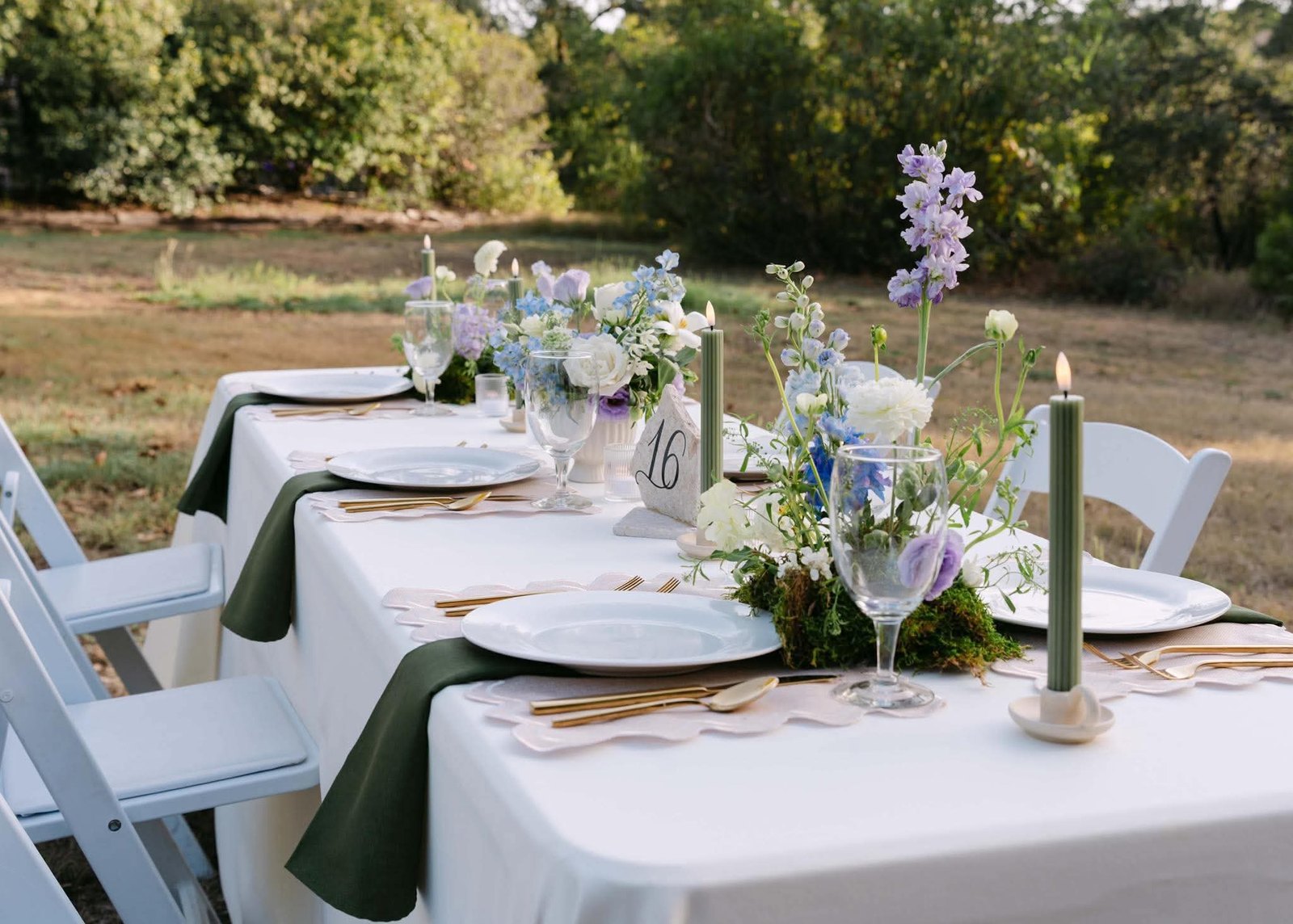 Reception table set in white linen with sage napkins, gold flatware, moss-set blue and lavender florals and green taper candles