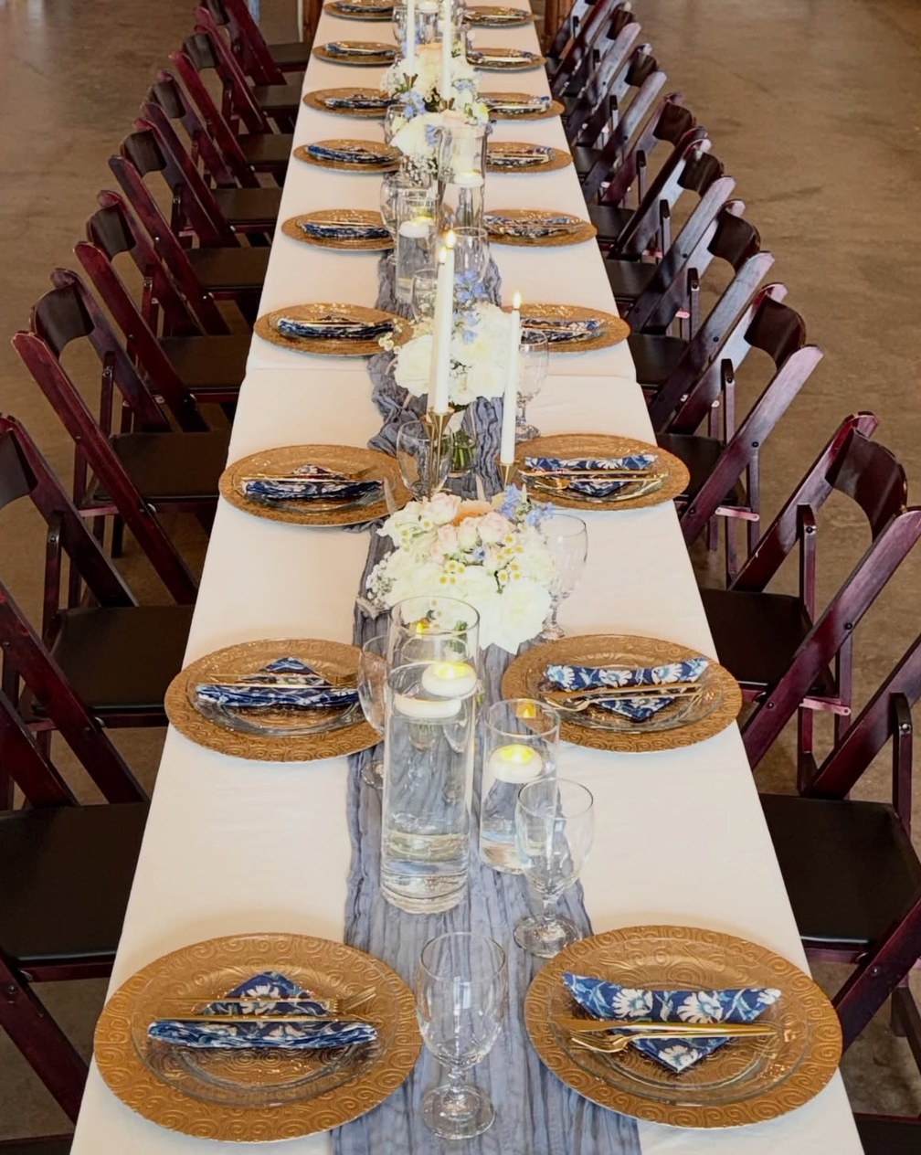 Long banquet table with gold chargers, blue toile napkins, white florals and taper candles