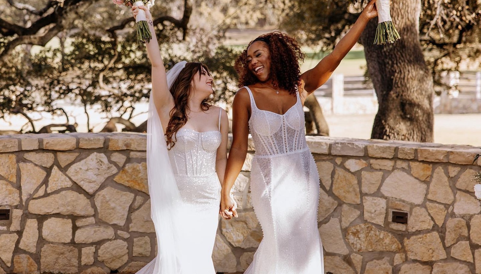 Two brides in white gowns laughing and holding bouquets overhead on a sunlit stone patio